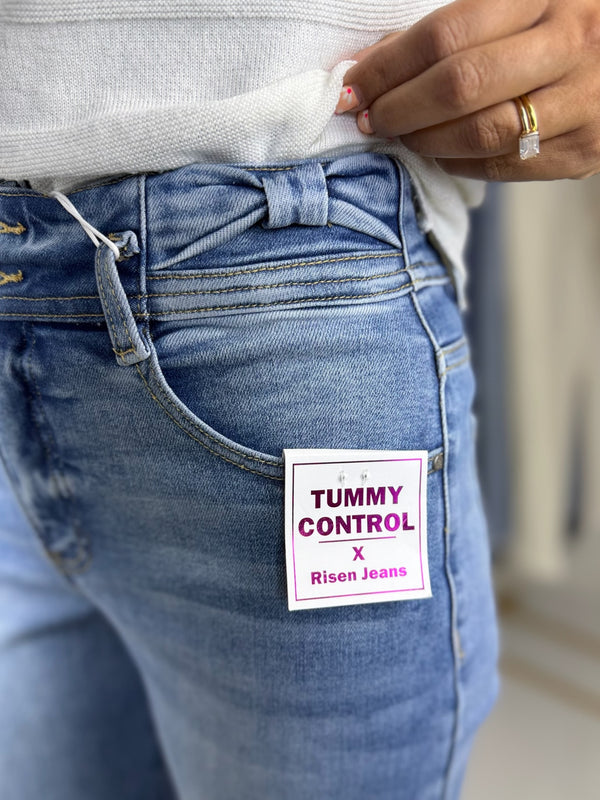 Close up of Blue jeans with a 'Tummy Control' label on the pocket and bow detail on waistband, worn by a person.