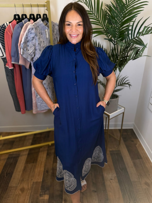 Woman wearing a blue dress standing in a room with a rack of clothes and a plant.