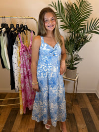 Woman wearing a blue floral dress standing in a room with a rack of clothes and a plant.