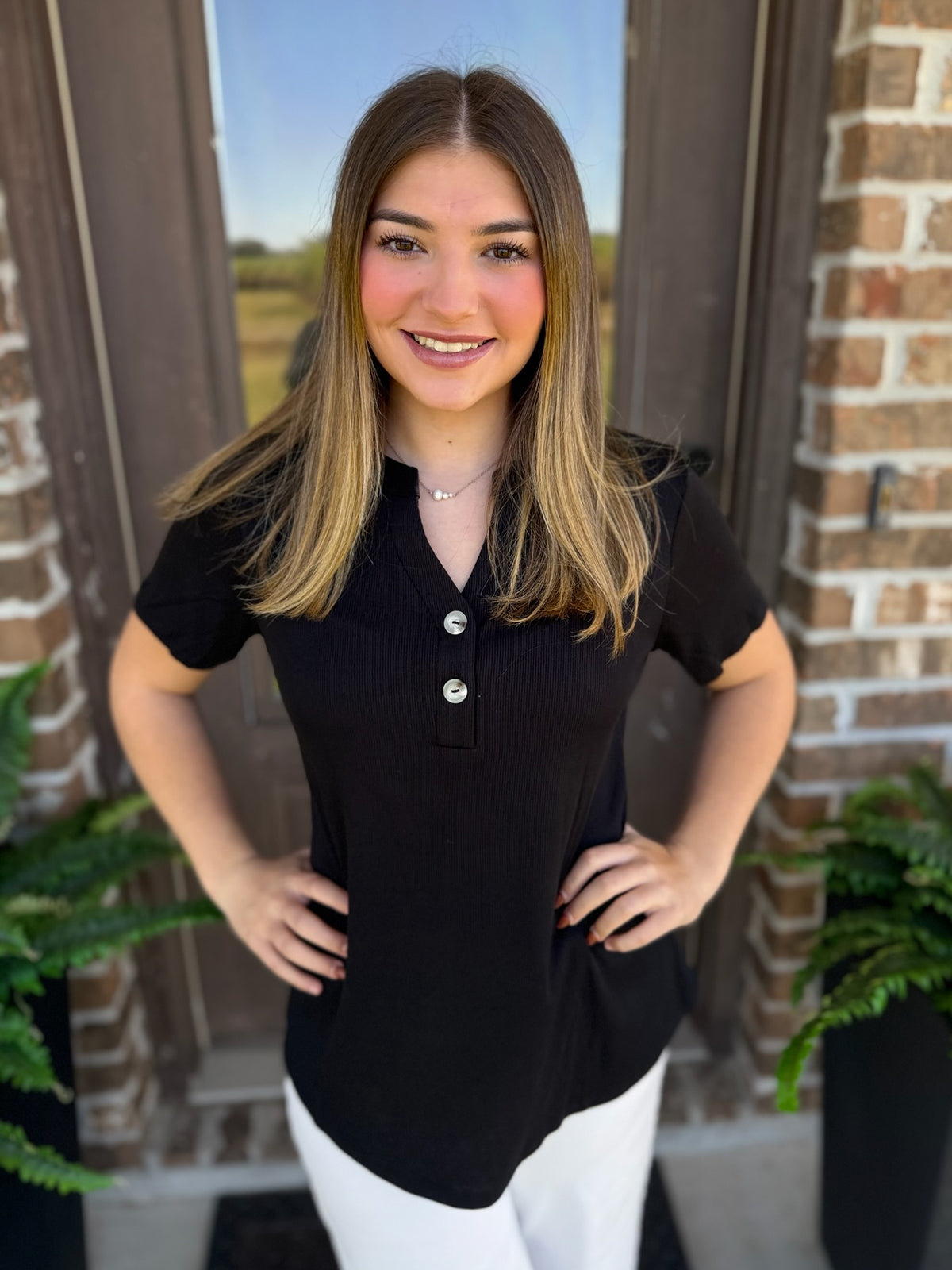 A woman with long brown hair stands in front of a brick door wearing a black short-sleeve henley shirt with white pants.