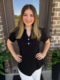 A woman with long brown hair stands in front of a brick door wearing a black short-sleeve henley shirt with white pants.