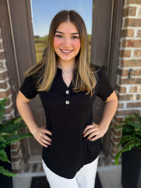 A woman with long brown hair stands in front of a brick door wearing a black short-sleeve henley shirt with white pants.