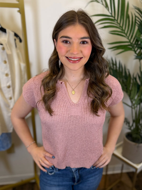 Woman wearing a pink knitted top with a plant and clothing rack in the background