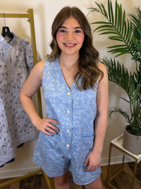 Woman wearing a blue floral v neck romper standing in a room with clothing and plants.