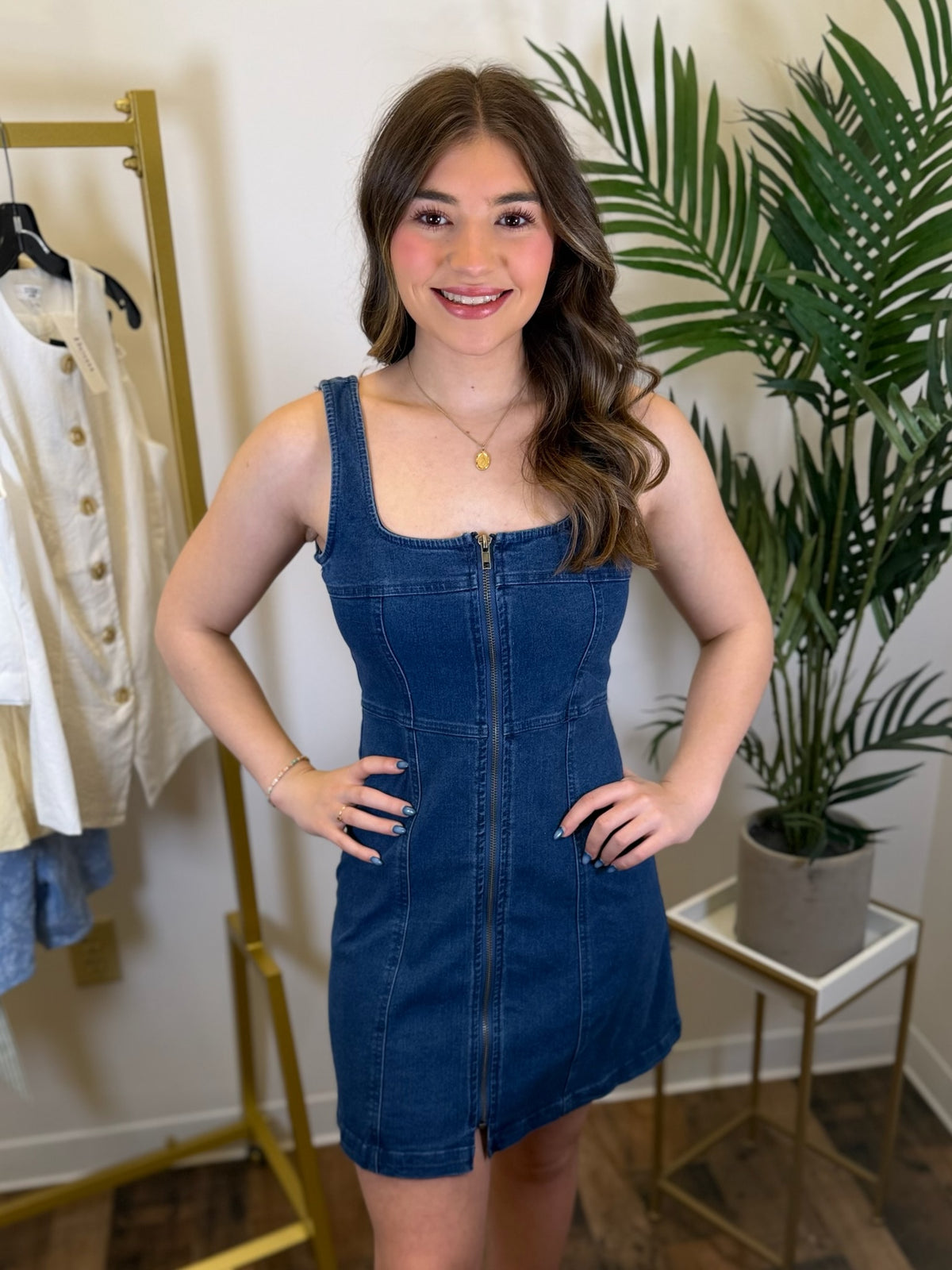 Woman wearing a blue denim dress standing in a room with plants and a rack of clothes.