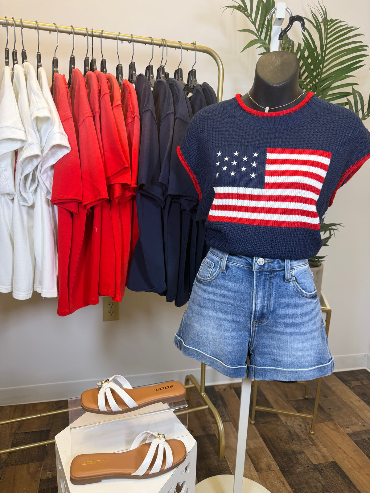 Mannequin wearing a navy sweater with an American flag design and denim shorts, surrounded by hanging clothes and shoes.