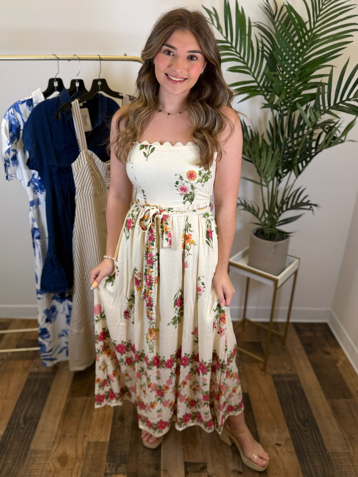 Woman wearing a sleeveless floral midi dress standing in a room with a clothing rack and plant.