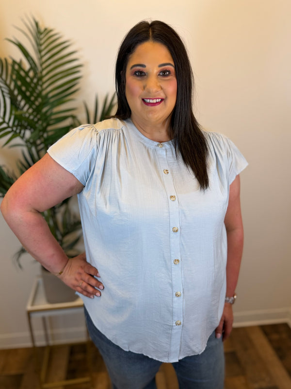 Woman wearing a light blue blouse with a white background and plant in the corner
