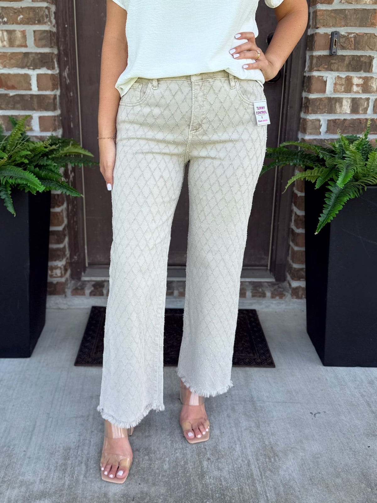A person modeling a pair of light beige textured wide-leg pants with a subtle diamond pattern and frayed hems, paired with a white top and clear high-heeled sandals, standing on a concrete porch in front of a brick door.