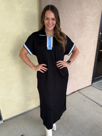 A woman modeling a black short-sleeve midi dress with light blue and white striped trim on the collar and sleeve cuffs, paired with white ankle boots, standing against a beige stucco wall.