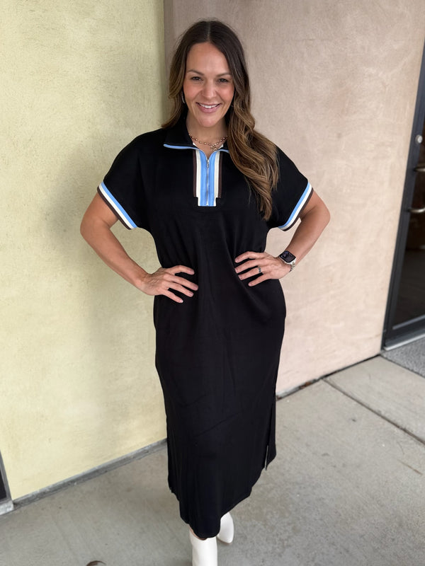 A woman modeling a black short-sleeve midi dress with light blue and white striped trim on the collar and sleeve cuffs, paired with white ankle boots, standing against a beige stucco wall.