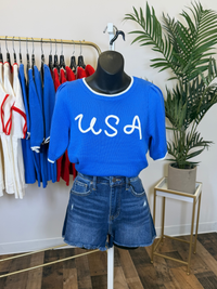 Blue short puff sleeve sweater top with 'USA' text and denim shorts on a mannequin in a store setting.