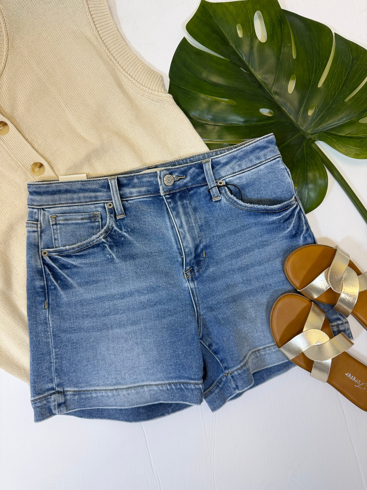 Blue denim shorts with a beige top and sandals on a white surface with a leafy plant.