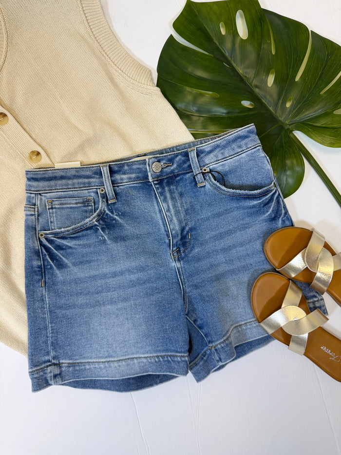 Blue denim shorts with a beige top and sandals on a white surface with a leafy plant.