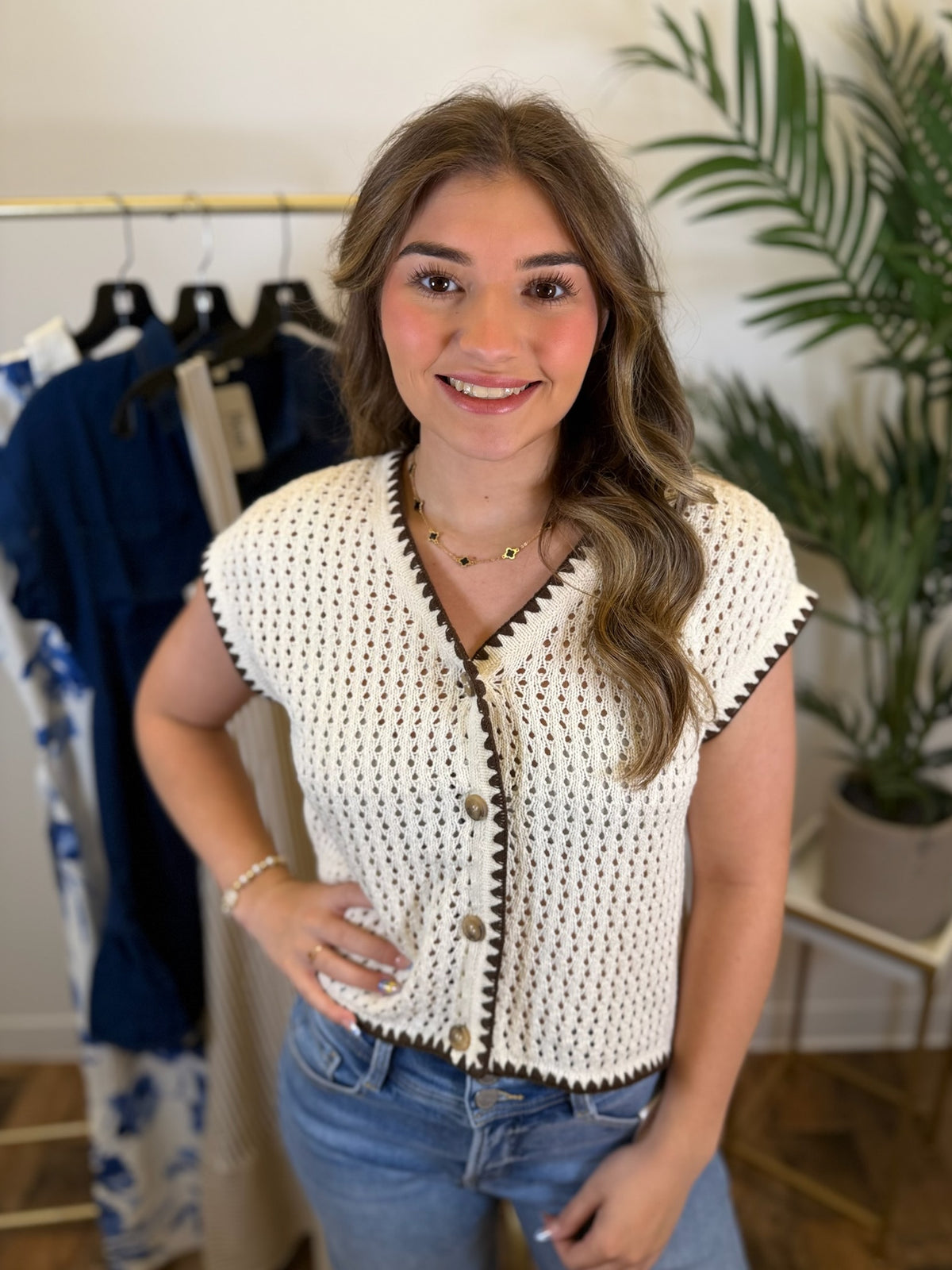 Woman wearing an off white short sleeve crochet top with button front and contrast trim details standing in a room with rack of clothing.