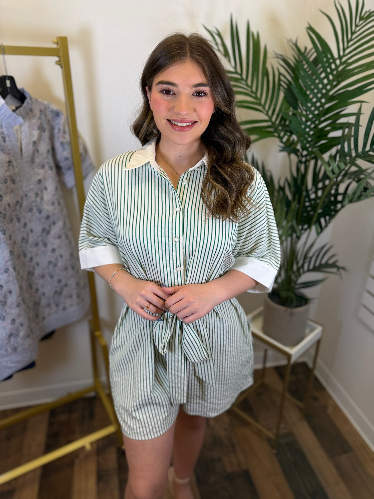 Woman wearing a green and white striped romper with white buttons and collar standing in a room with plants and clothing racks.