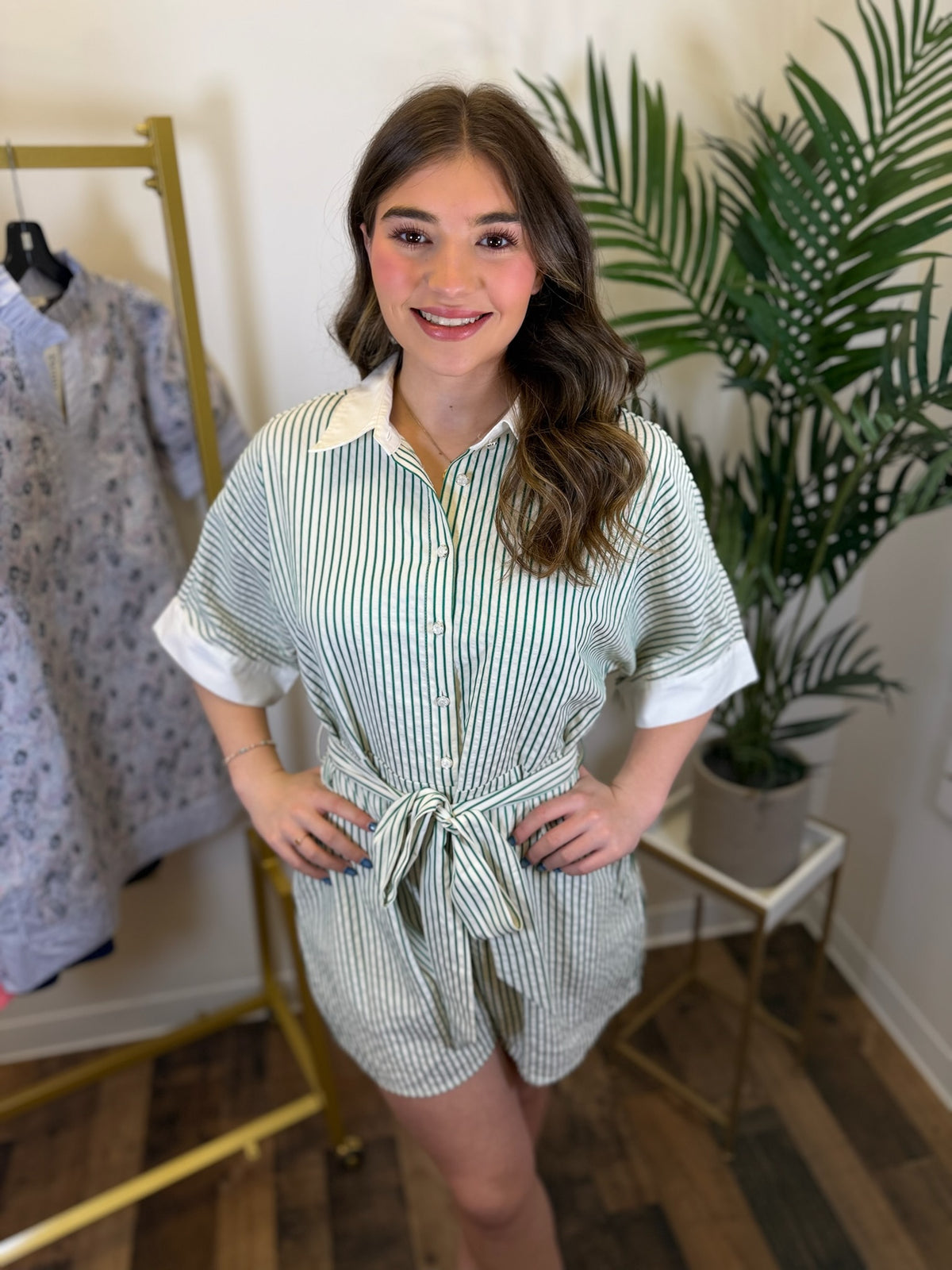 Woman wearing a striped romper with button front and tie waist in a clothing store with plants in the background