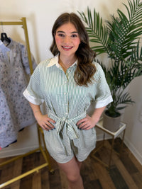 Woman wearing a striped romper with button front and tie waist in a clothing store with plants in the background