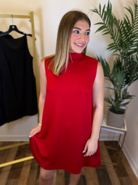 A woman wearing a sleeveless, high-neck red dress, standing in a room with a clothing rack, potted plant, and wooden floor.