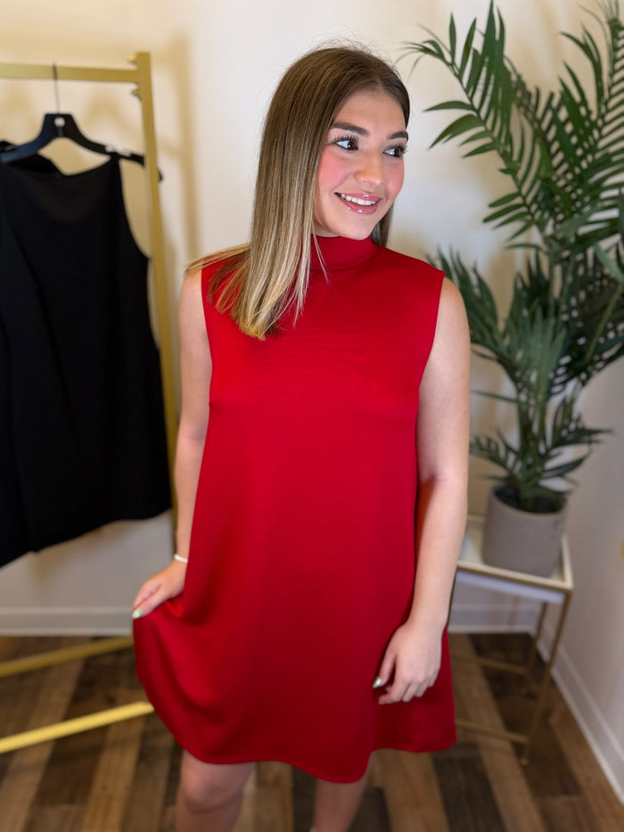 A woman wearing a sleeveless, high-neck red dress, standing in a room with a clothing rack, potted plant, and wooden floor.