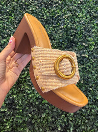 Hand holding a open toe heel with a woven strap and gold ring on a grassy background