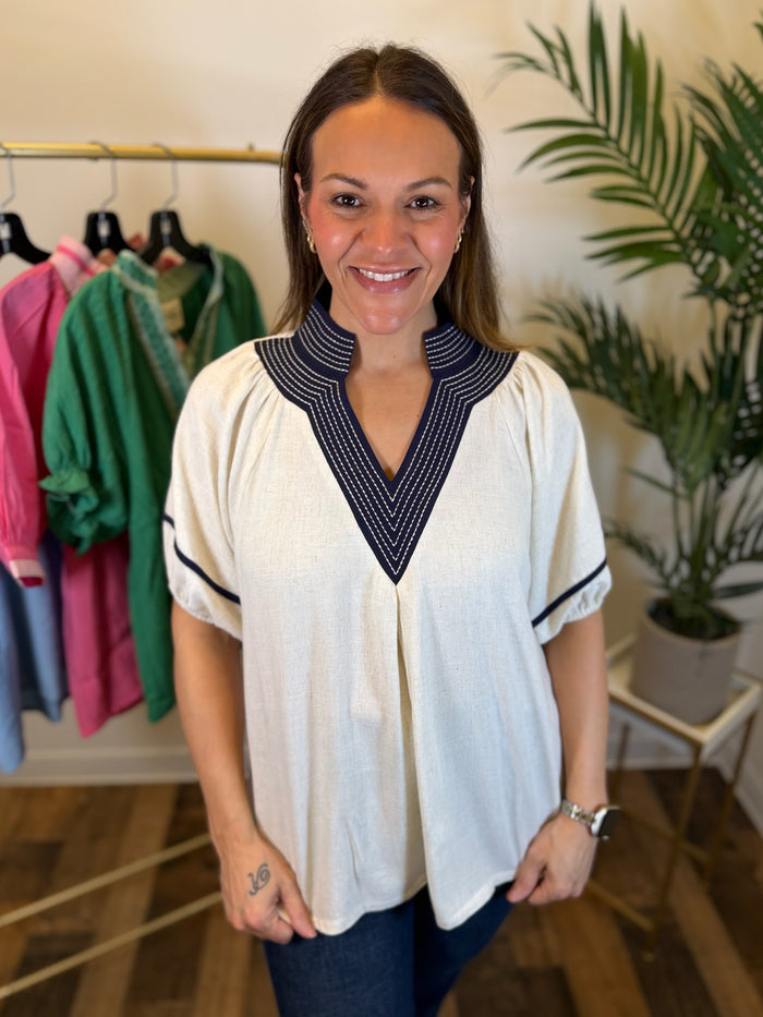 A woman smiling while wearing a cream-colored short-sleeve blouse with a navy blue and white patterned V-neck collar and sleeve trim, standing in a boutique setting with clothing racks and a potted plant in the background.