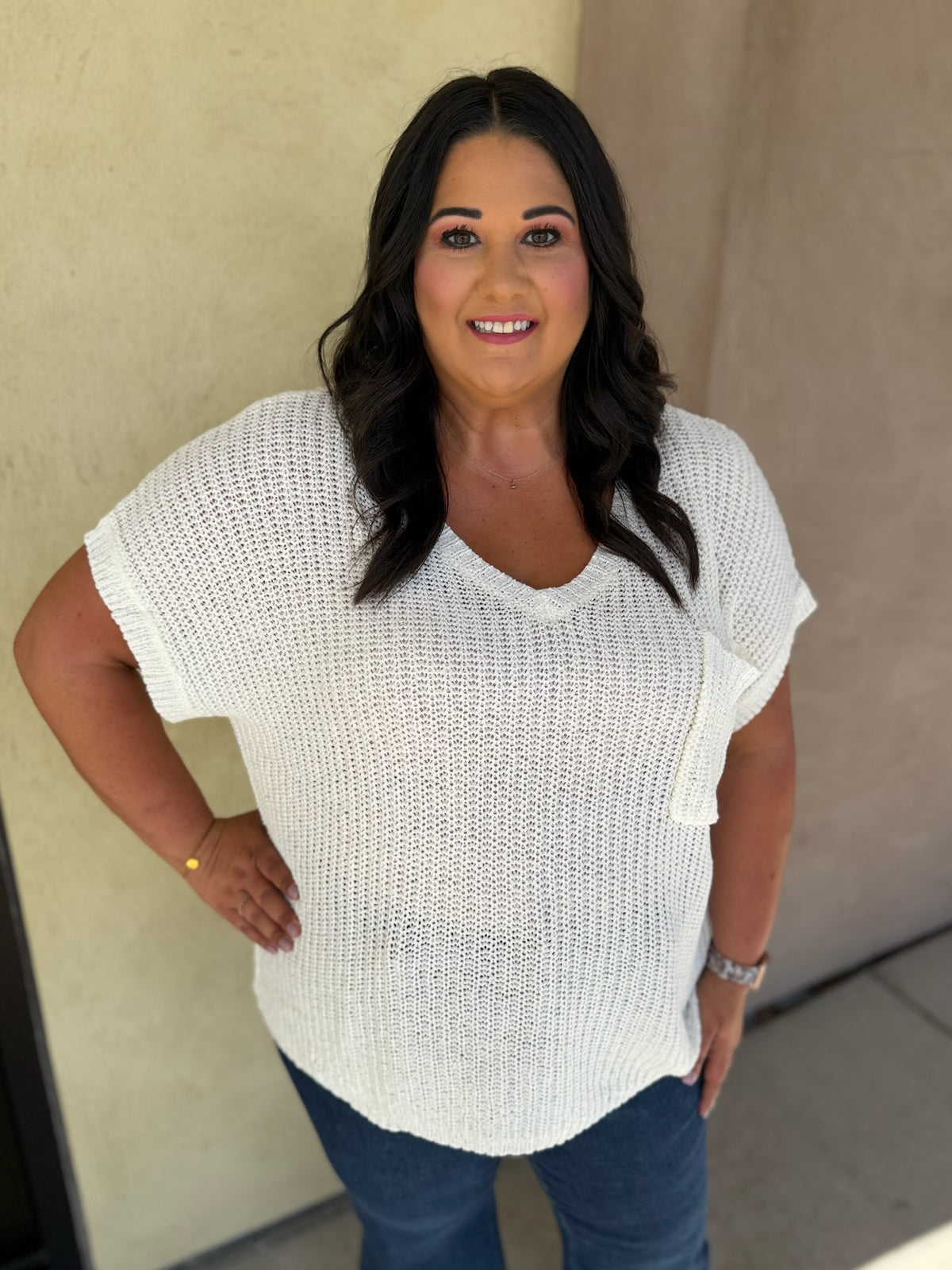 A woman wearing a white textured knit short-sleeve V-neck top and blue jeans, standing against a beige stucco wall.