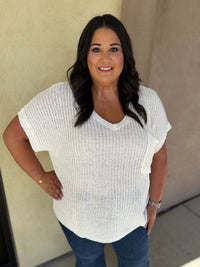 A woman wearing a white textured knit short-sleeve V-neck top and blue jeans, standing against a beige stucco wall.