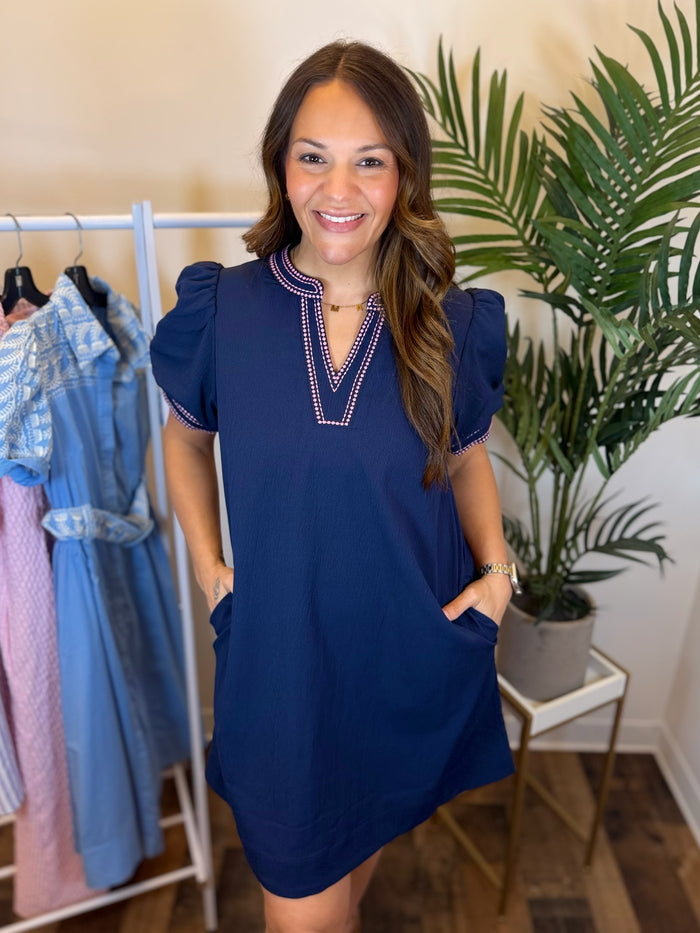 Woman wearing Navy mini dress with short puff sleeves, split neckline, and pink trim details in a clothing store setting.