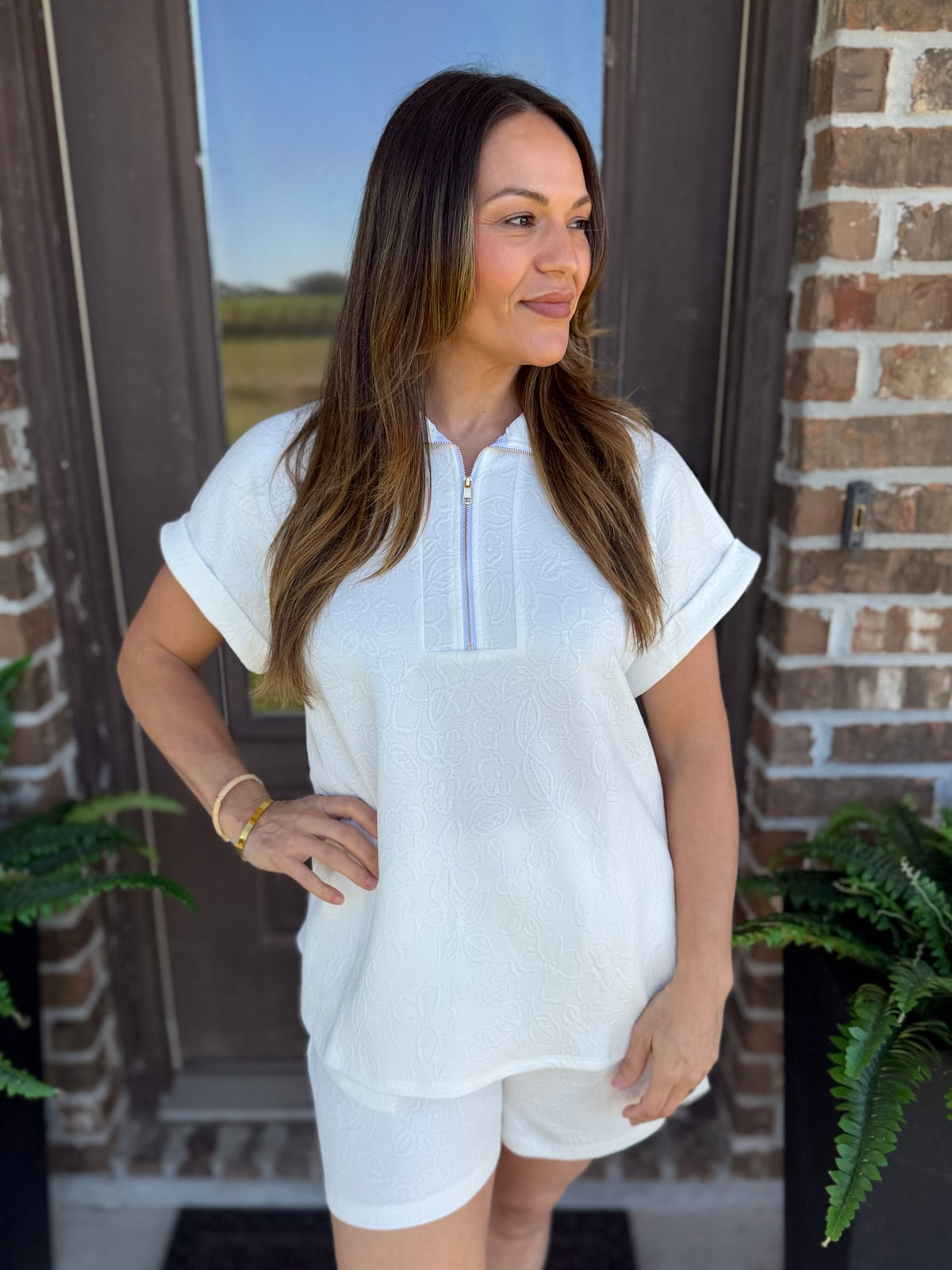 A woman modeling a white short-sleeve zip-front top and matching white shorts with flower texture, standing in front of a brick door with plants visible on both sides.