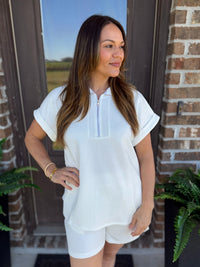 A woman modeling a white short-sleeve zip-front top and matching white shorts with flower texture, standing in front of a brick door with plants visible on both sides.
