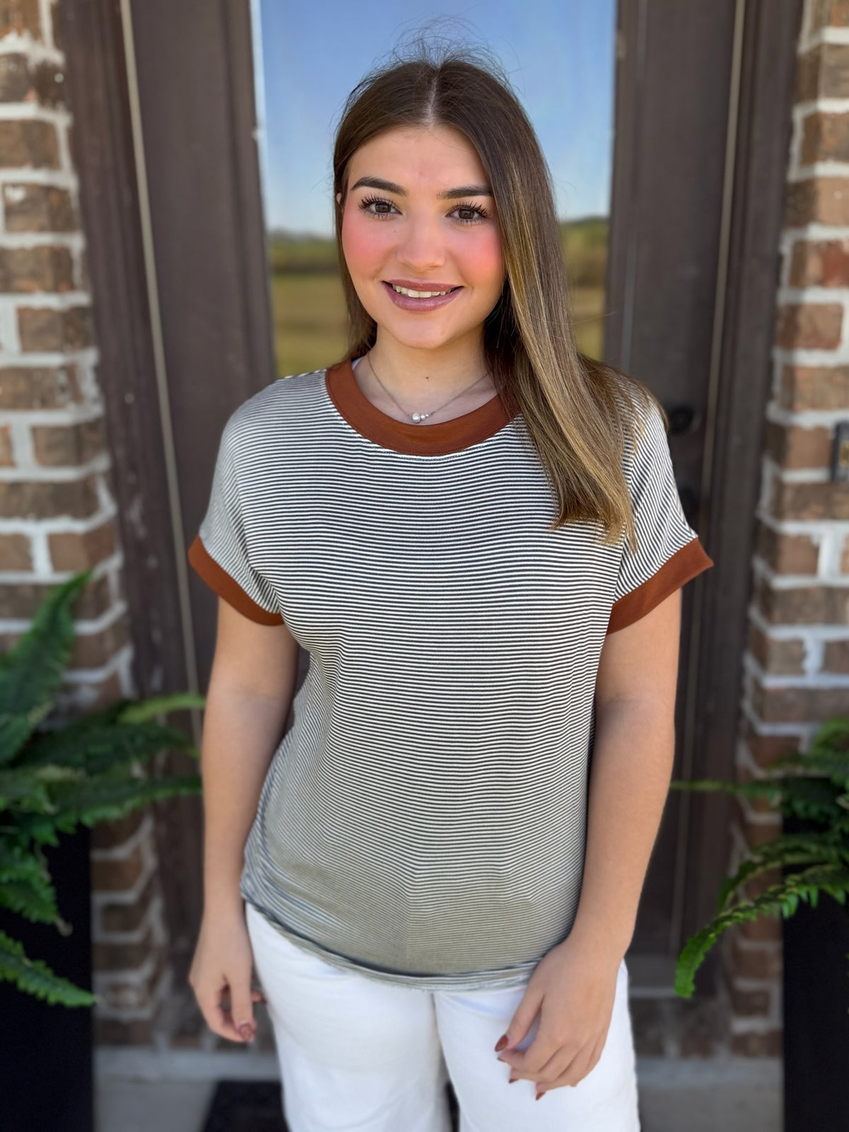A woman wearing a short-sleeve ringer t-shirt with fine horizontal olive and white stripes and contrasting brown collar and sleeve cuffs, standing in front of a brick door.