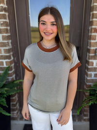 A woman wearing a short-sleeve ringer t-shirt with fine horizontal olive and white stripes and contrasting brown collar and sleeve cuffs, standing in front of a brick door.