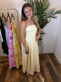 Woman in a light yellow strapless dress with lace trim standing in a room with a rack of dresses and a plant.