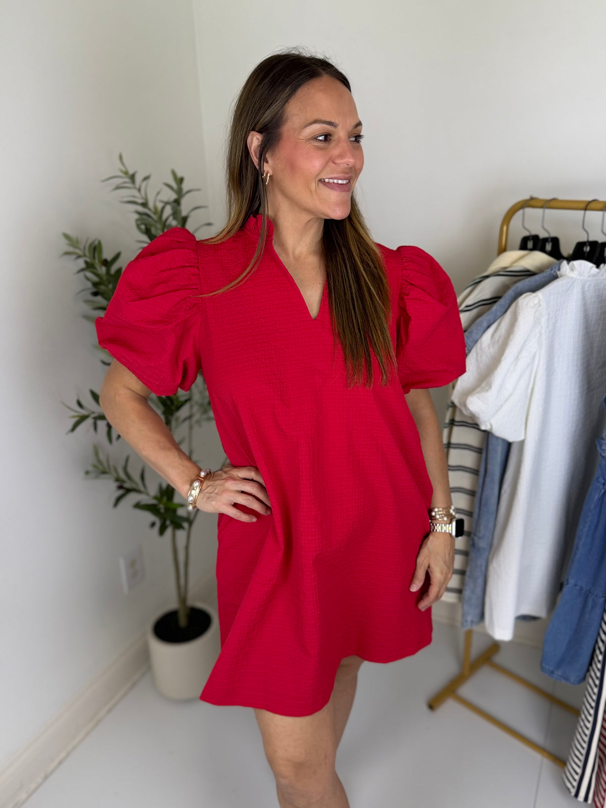 Woman wearing a red textured mini dress standing in a room with a plant and clothing rack.