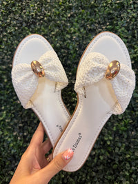 White sandals with decorative bows and gold hardware held against a green grass background