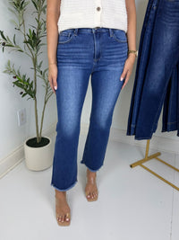 Person wearing blue jeans standing in a room with a plant and coat rack.