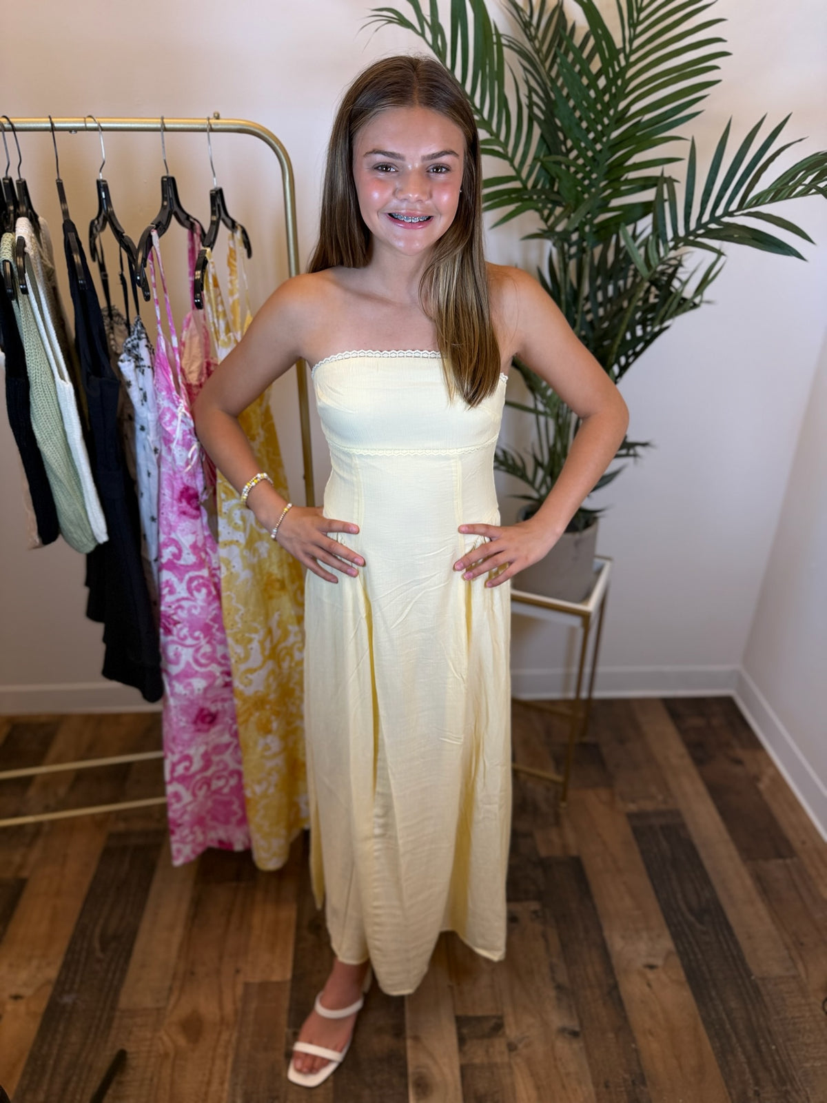Young girl in a strapless butter yellow midi dress with lace trim standing in a room with a rack of clothes and a plant.