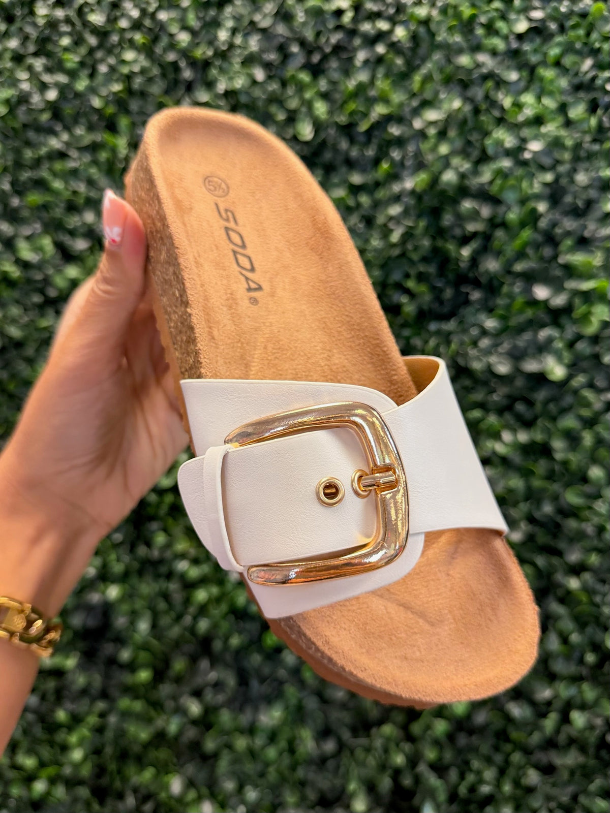 White slip on sandal with gold buckle held against a green textured background