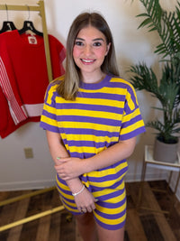 A woman wearing a matching yellow and purple striped t-shirt and shorts set, standing in a room with a clothing rack and a potted plant in the background.