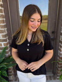 A woman wearing a black short-sleeve polo shirt with a three-button placket and white pants, standing in front of a brick door.