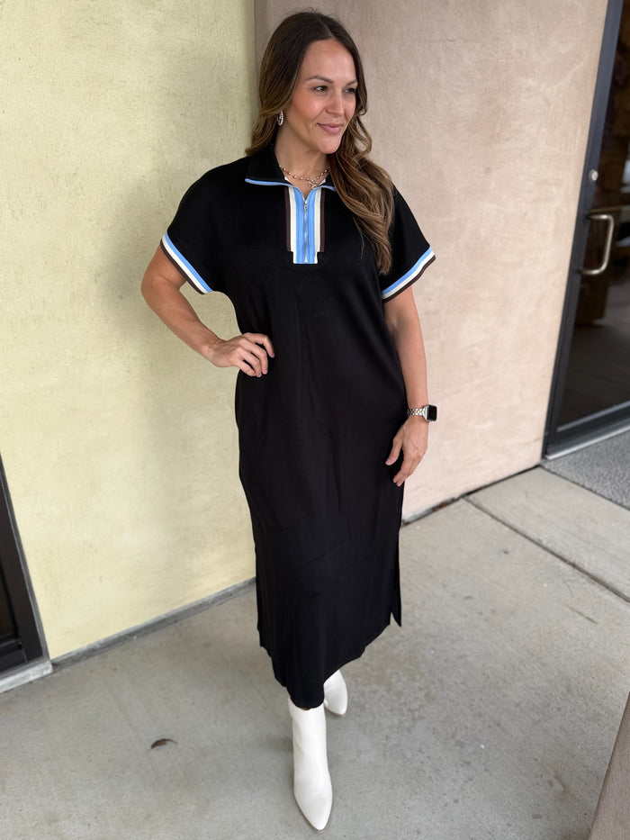 A woman modeling a black short-sleeve midi dress with light blue and white striped trim on the collar and sleeve cuffs, paired with white ankle boots, standing against a beige stucco wall.