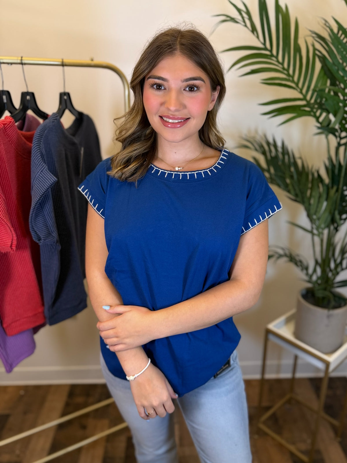 A woman wearing a royal blue short-sleeve top with white stitch trim around the neckline and sleeve cuffs, paired with light blue jeans, standing in a boutique setting with a clothing rack and a potted plant in the background.