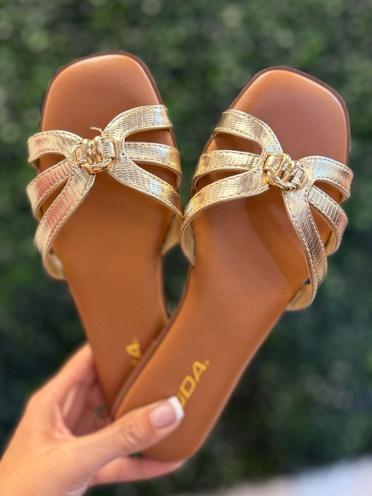 Gold sandals with decorative bows held by a hand against a blurred green background