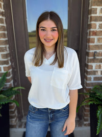 A woman wearing a white short-sleeve waffle knit textured top with a chest pocket and blue jeans, standing in front of a brick doorway with plants.
