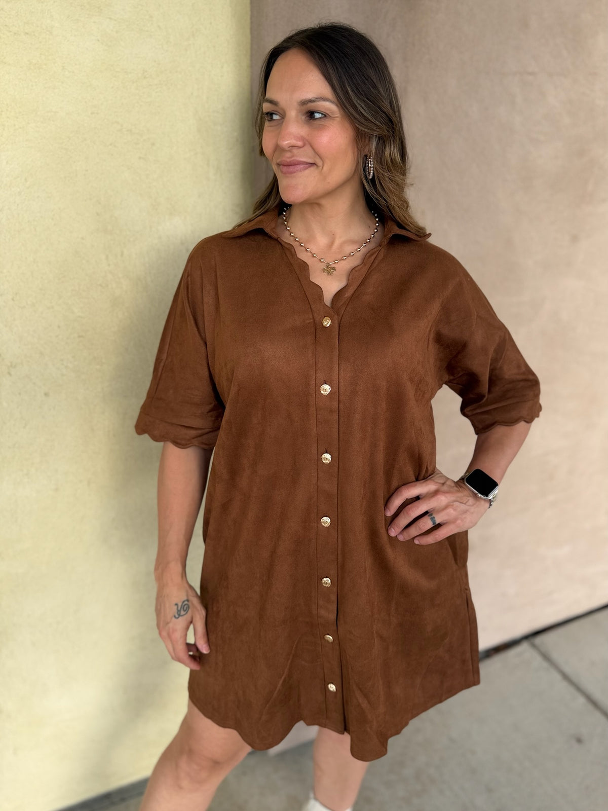 A woman wearing a short-sleeve camel button-front shirt dress with scalloped trim and pockets, standing outdoors against a beige stucco wall.