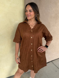 A woman wearing a short-sleeve camel button-front shirt dress with scalloped trim and pockets, standing outdoors against a beige stucco wall.