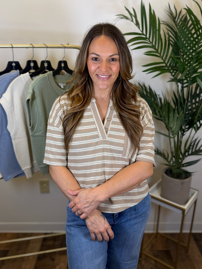 Woman standing in a room with clothing and plants wearing jeans and a tan and white striped short sleeve top with button neck and chest pocket.