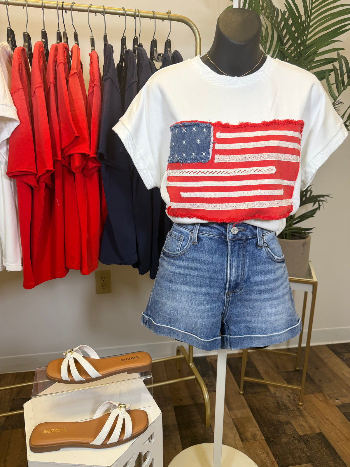 Mannequin wearing a white t-shirt with an American flag design and denim shorts, with red and blue shirts hanging in the background.