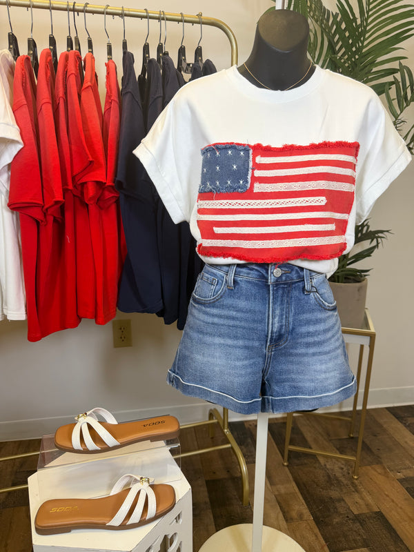 Mannequin wearing a white t-shirt with an American flag design and denim shorts, with red and blue shirts hanging in the background.